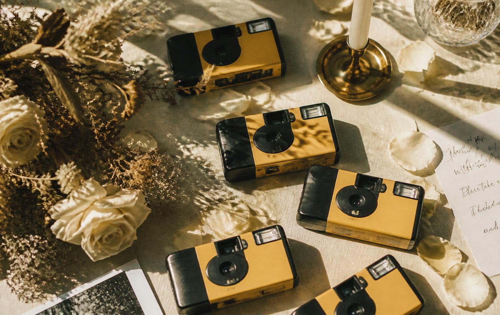 A vintage disposable film camera resting on ivory marble beside a printed cream QR card with gold foil border and eucalyptus